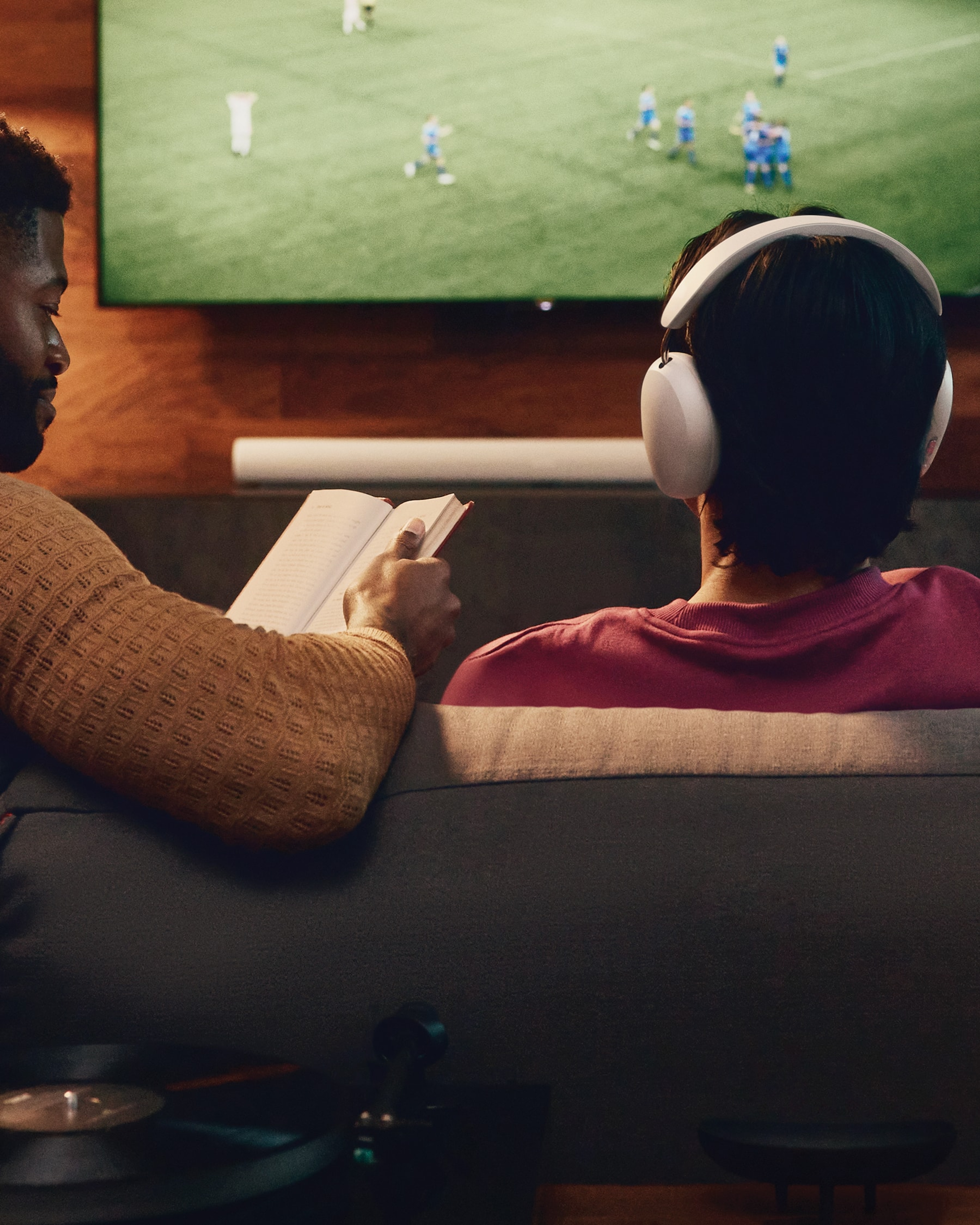 Two people watching TV; one reading, the other wearing white Sonos Ace headphones