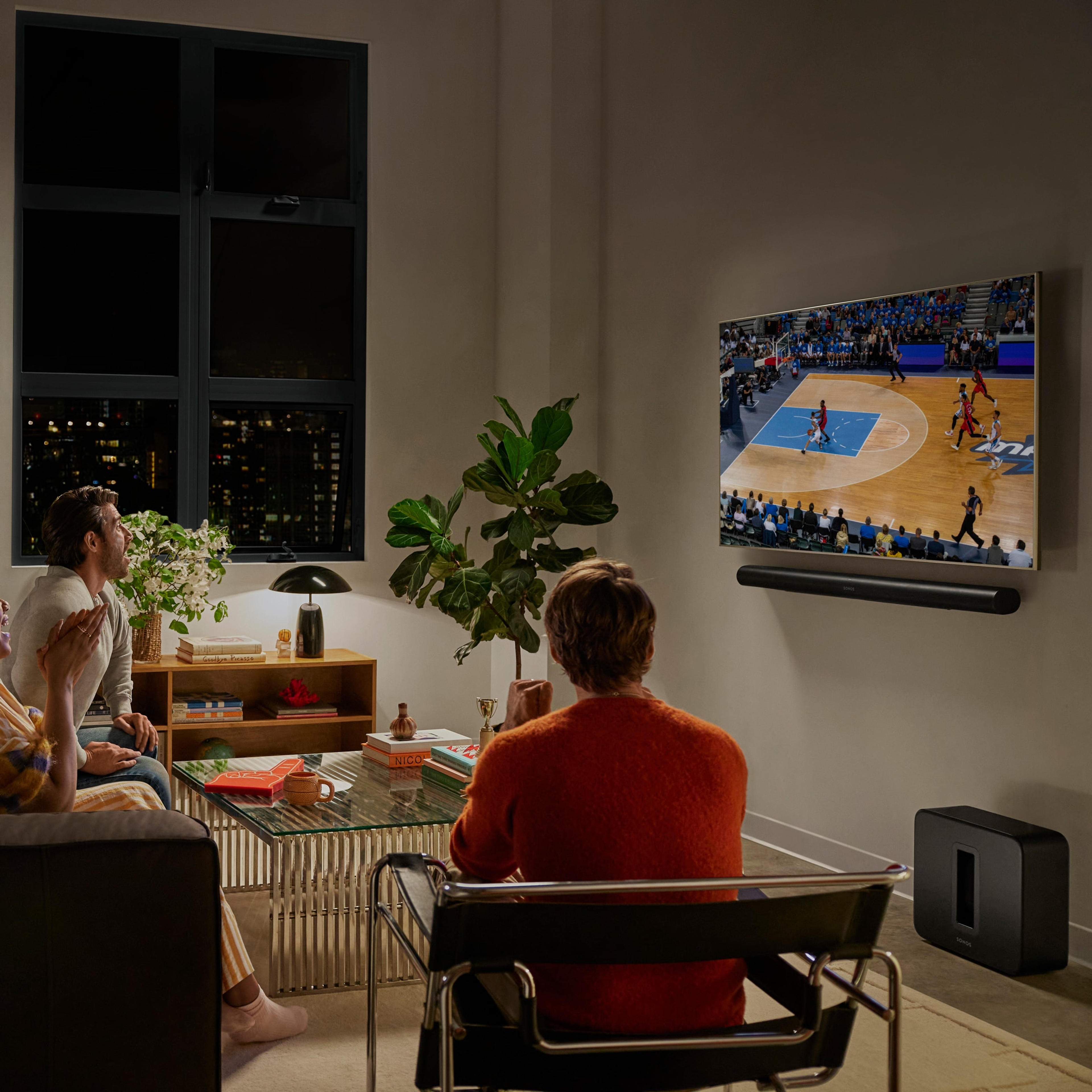 En grupp vänner som tittar på en basketmatch i ett vardagsrum med en svart Arc Ultra och en svart Sub 4